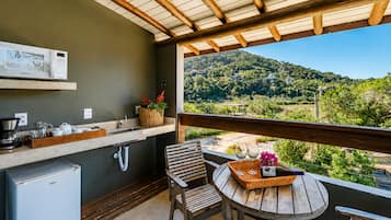 A balcony with a wooden table, a chair, a small refrigerator, a sink, and a view of a green landscape.