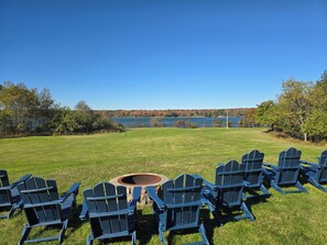 A grassy outdoor area with a fire pit and several Adirondack chairs arranged facing a lake. - Oceanfront house close to Acadia National Park! (Ellsworth)