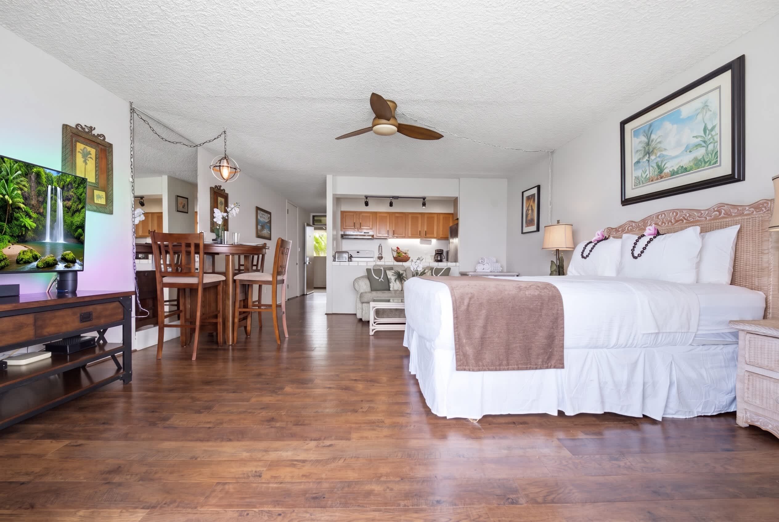Een ruime kamer met een groot bed, een zithoek met stoelen, een keuken achtergrond en een plafondventilator.