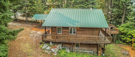 A cabin with a green roof, wooden deck, and a stone foundation.