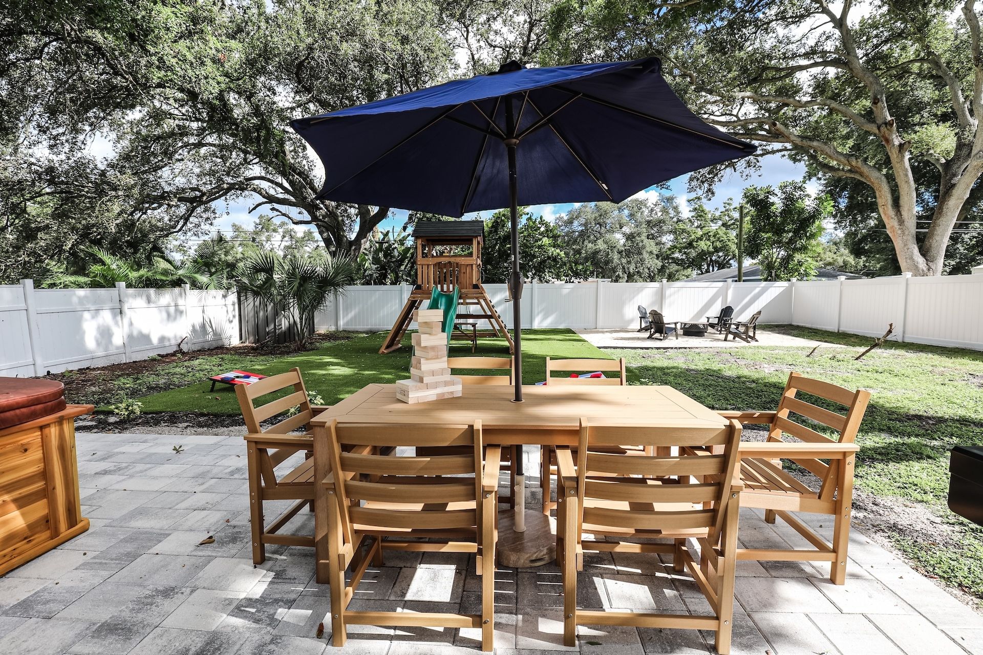 Un espace de restauration en plein air avec une table et des chaises en bois, un parasol bleu et, au fond, une structure de jeux pour enfants.