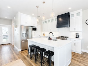 A modern kitchen with white cabinetry, a stainless steel refrigerator, a gas stove, and a large island with bar stools. - Jamaica Beach | Bayfront | Hot Tub | Kayaks (Jamaica Beach)