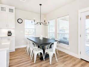 A dining area with a dark wooden table, white chairs, a chandelier, and large windows with blinds. - Jamaica Beach | Bayfront | Hot Tub | Kayaks (Jamaica Beach)