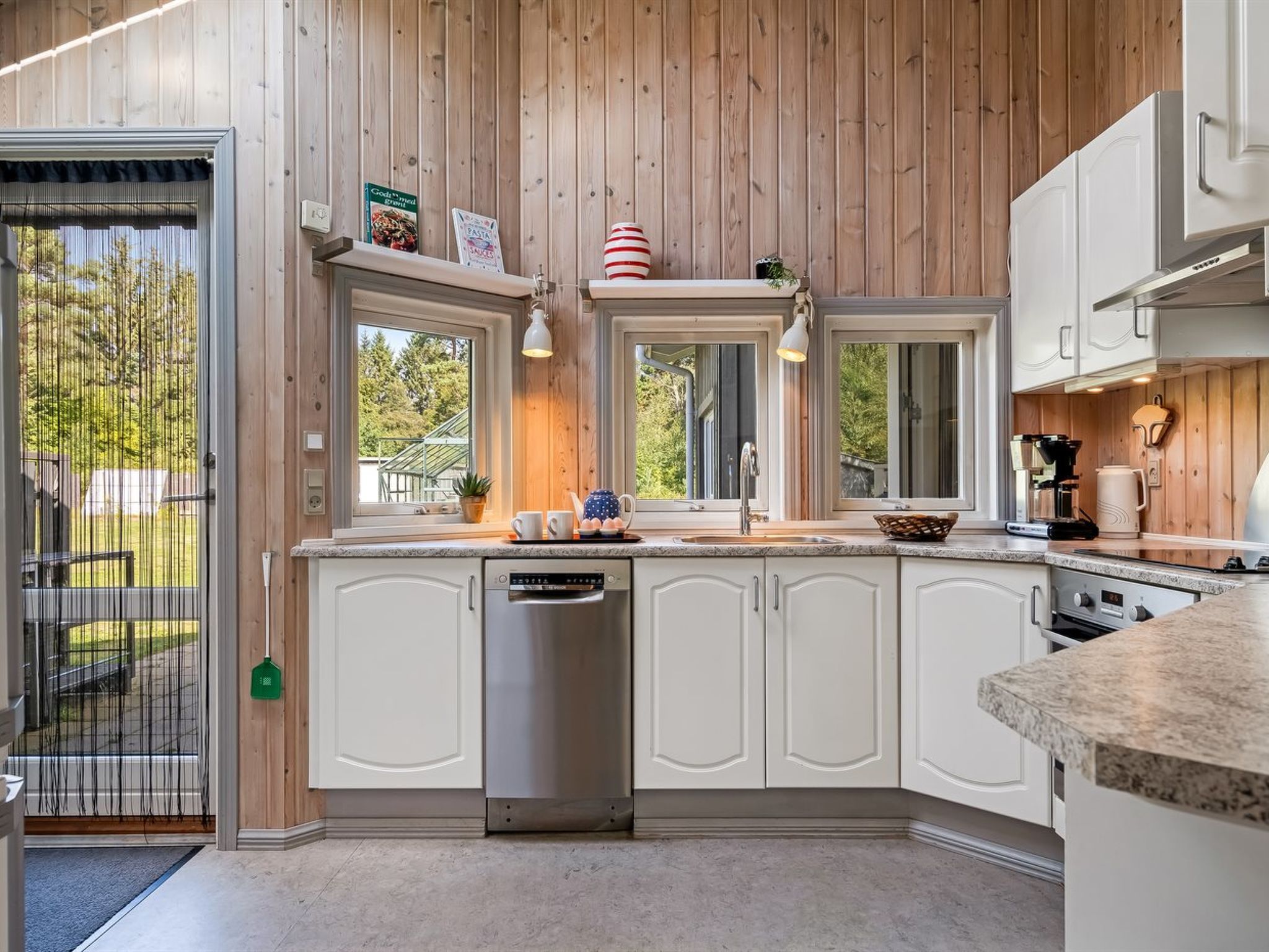 A modern kitchen with wooden paneling, white cabinetry, a built-in dishwasher, and a large window.