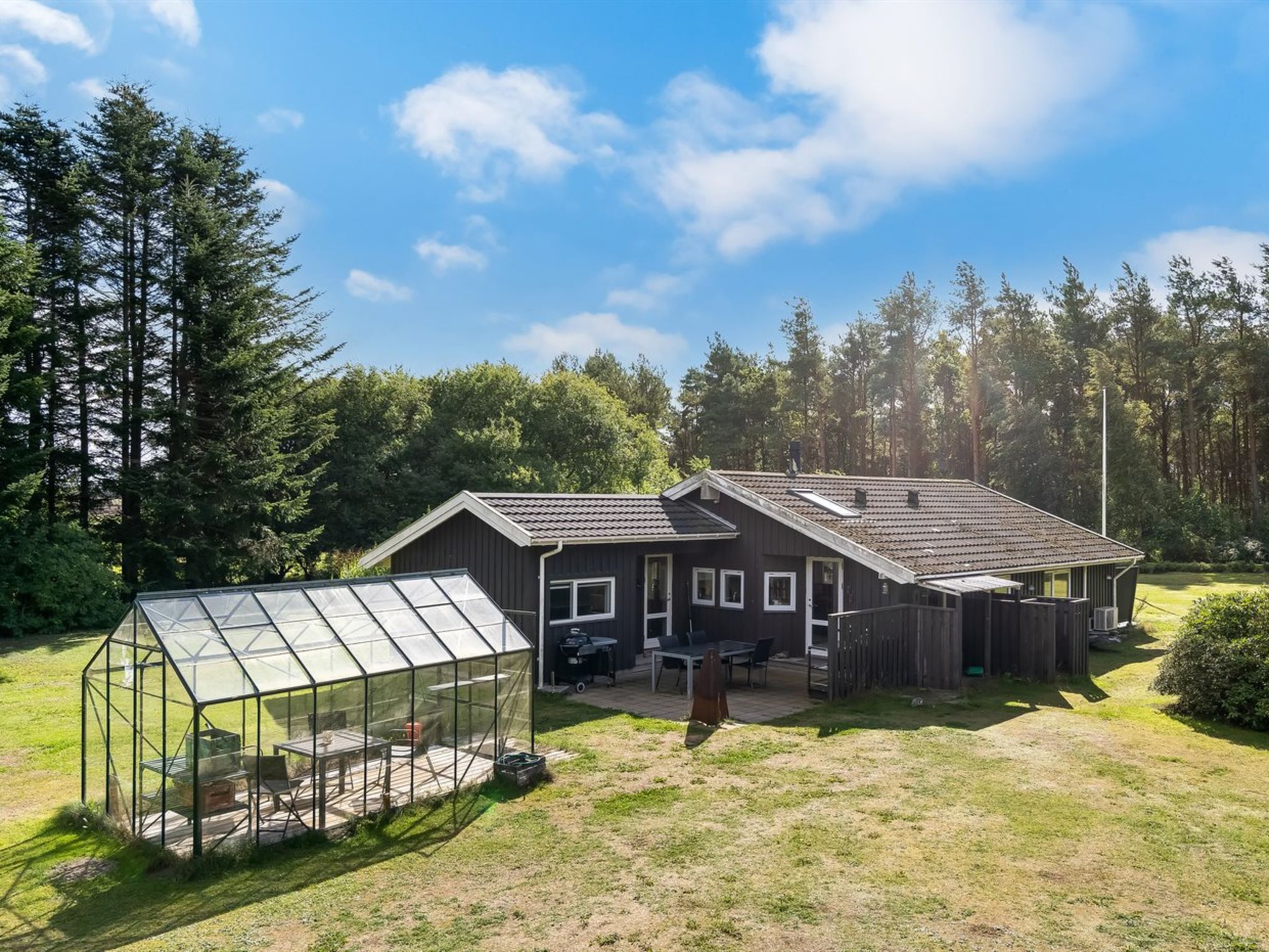 A two-story house with a large deck, a greenhouse, and a forested backdrop.