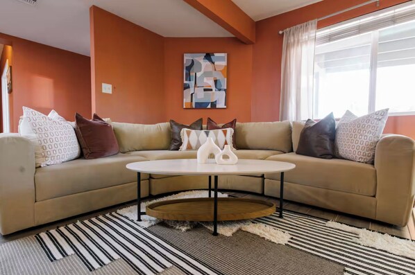 Una sala de estar moderna con un sofá seccional color beige, una mesa de centro redonda y un cuadro abstracto grande en la pared.