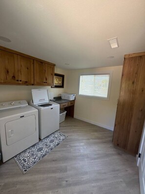 A laundry room with two washing machines, a dryer, a wooden cabinet, a window with blinds, and a patterned rug.