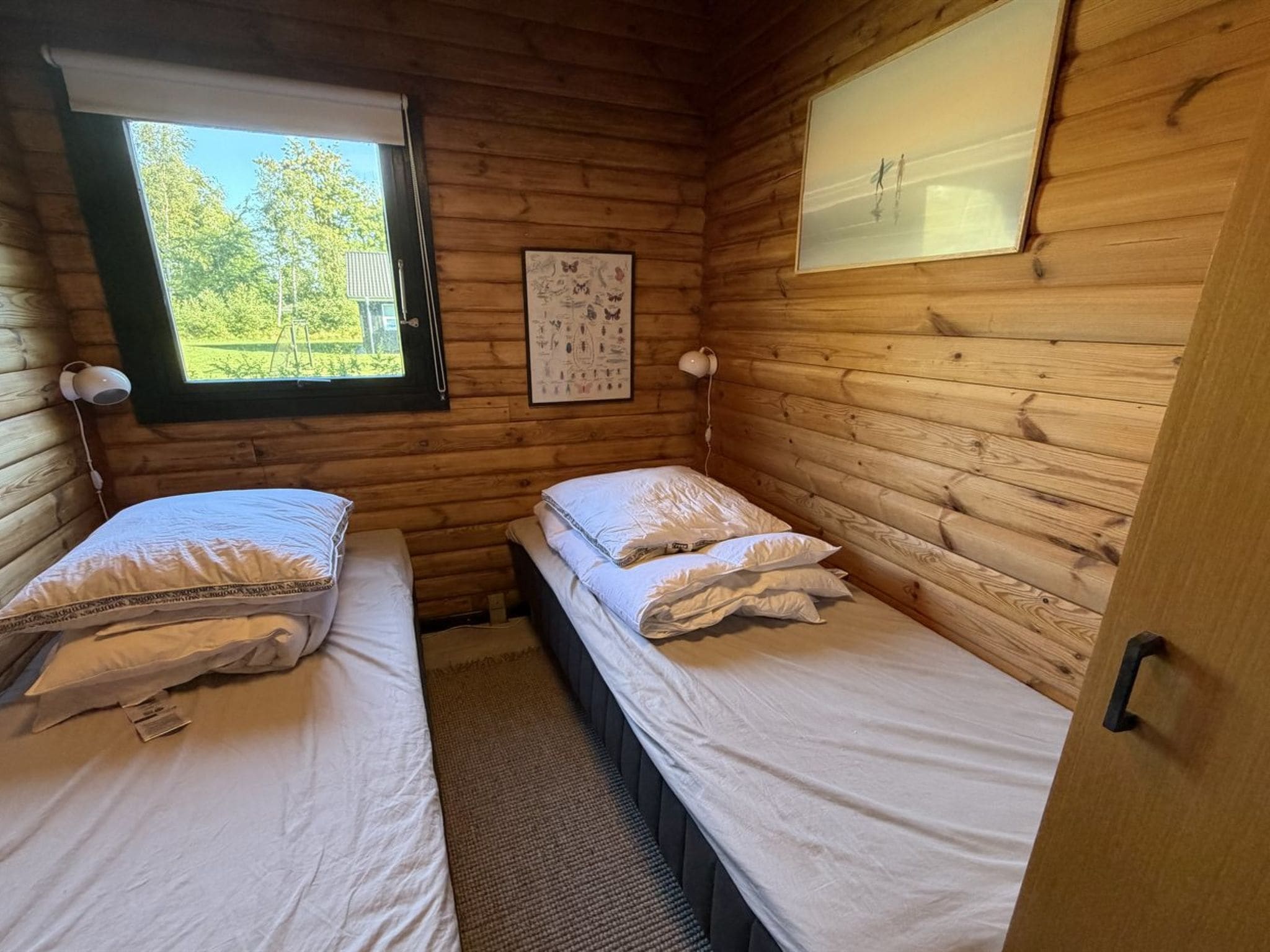 Een kamer in een blokhut met twee bedden, een raam, een wandlamp en een lijst met een afbeelding.