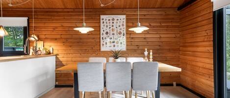 A modern kitchen with wooden walls, a dining table with four chairs, pendant lights, and a large framed picture on the wall.