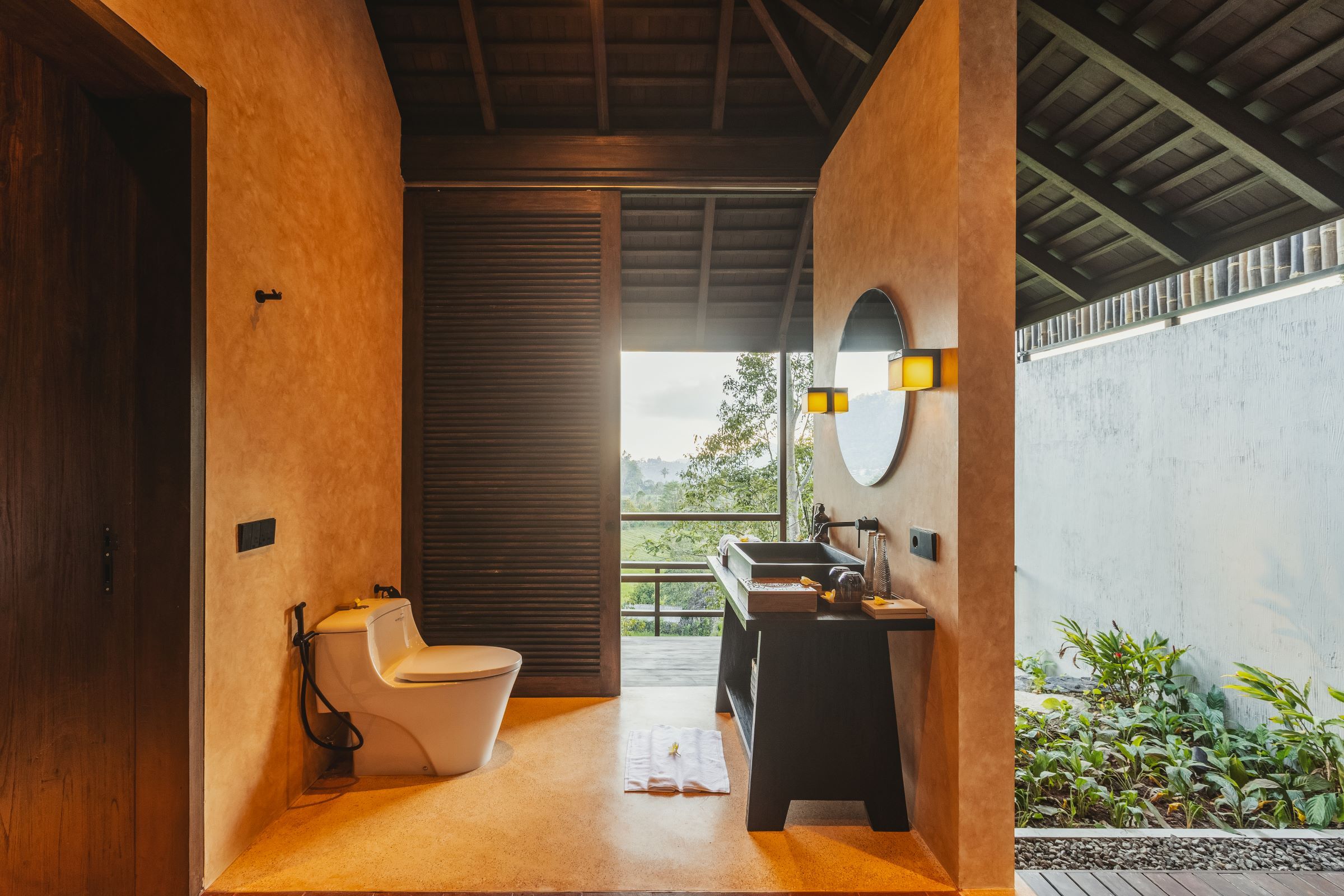 A modern bathroom with a toilet, sink, and a view of greenery outside.