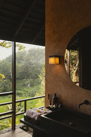 Un baño con vista a un paisaje exuberante, un espejo redondo y un lavamanos con artículos de tocador.