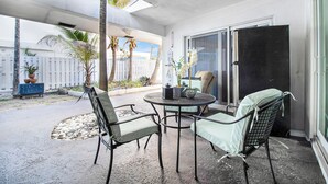 Un patio al aire libre con una mesa y sillas, un ventilador de techo y vistas del entorno, incluyendo palmeras y una cerca blanca.
