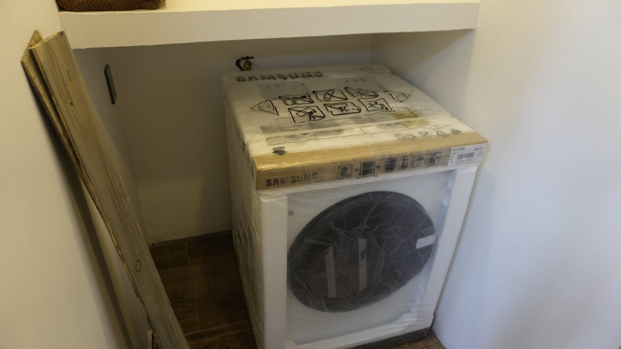 Une petite machine à laver blanche, placée dans un coin d’une pièce dont le sol est en bois.