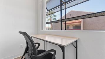Une pièce avec un bureau, une chaise, une fenêtre donnant sur des immeubles et des palmiers.