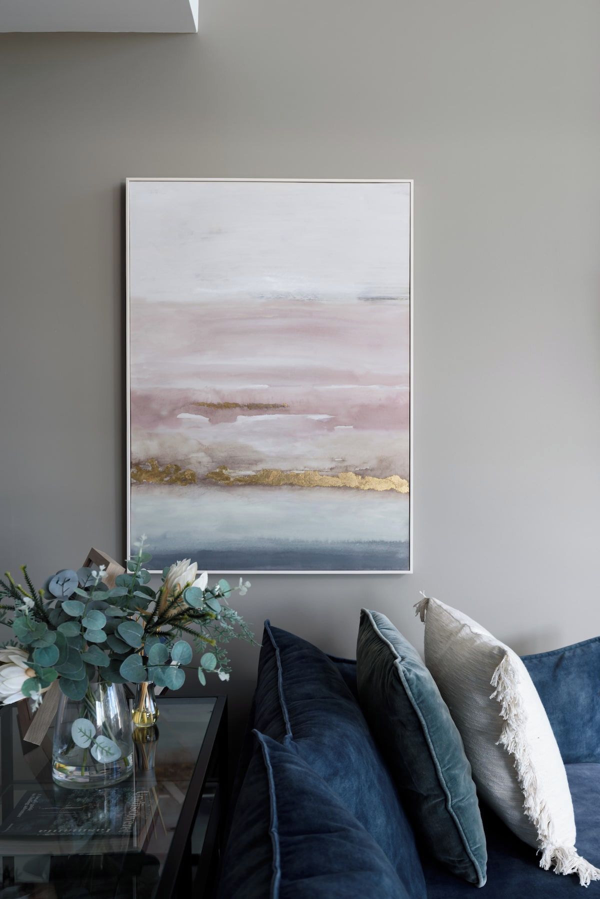 A room with a large abstract painting on the wall, a glass table with a vase of greenery, and a blue sofa with cushions.