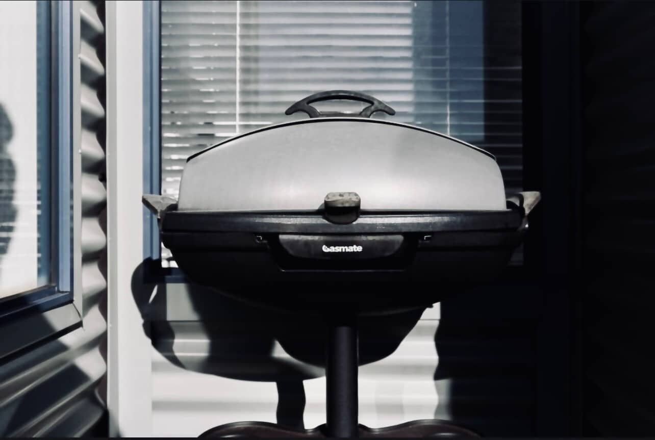 Una parrilla de gas con un asa, ubicada frente a una ventana con persianas.