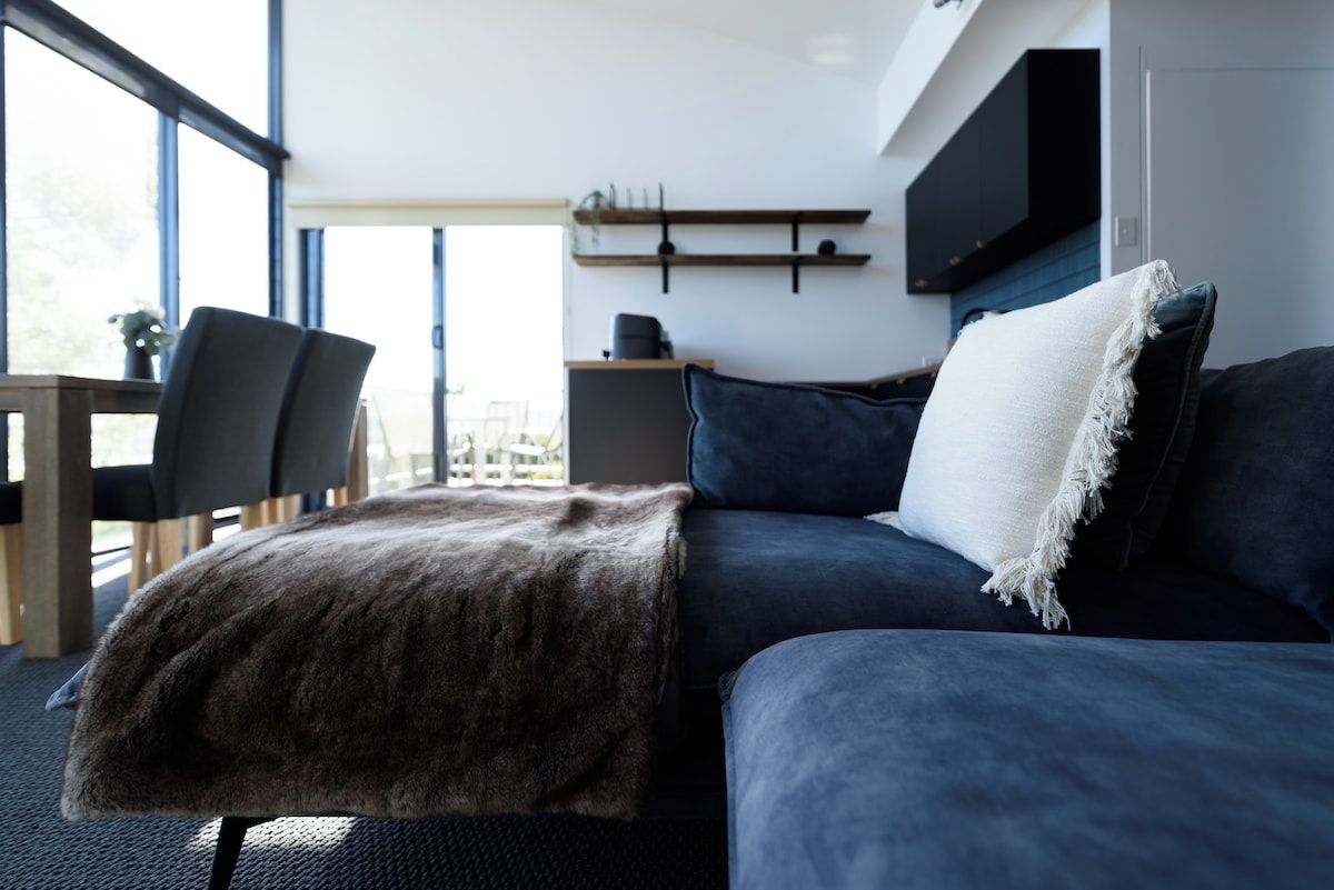 Un salon moderne avec un canapé bleu foncé, un coussin blanc et un plaid en fausse fourrure. On aperçoit un meuble bas en bois et une chaise au premier plan.