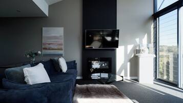 A modern living room with a dark blue sofa, a glass coffee table, and a flat-screen TV mounted on a dark wall.