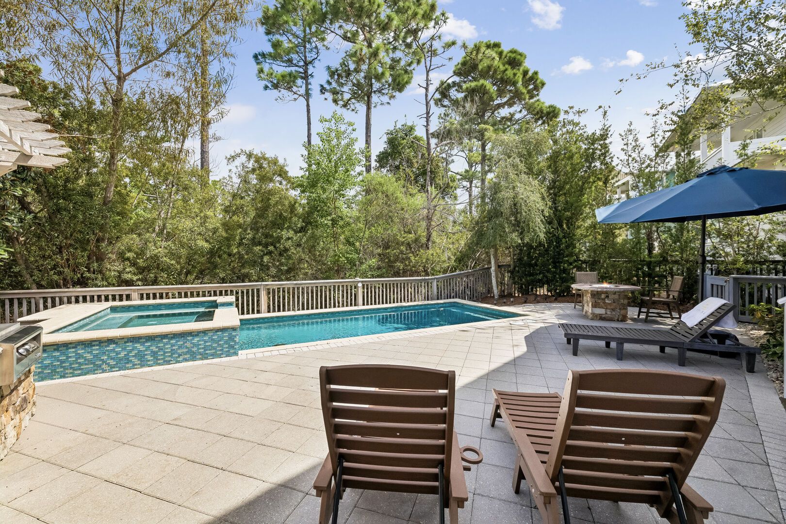 A swimming pool with a wooden deck, lounge chairs, and a stone fire pit.