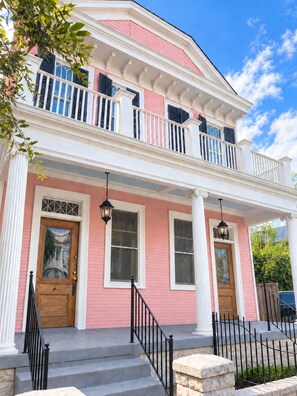 Una casa de dos pisos con fachada rosa, detalles blancos y un balcón con barandillas negras.