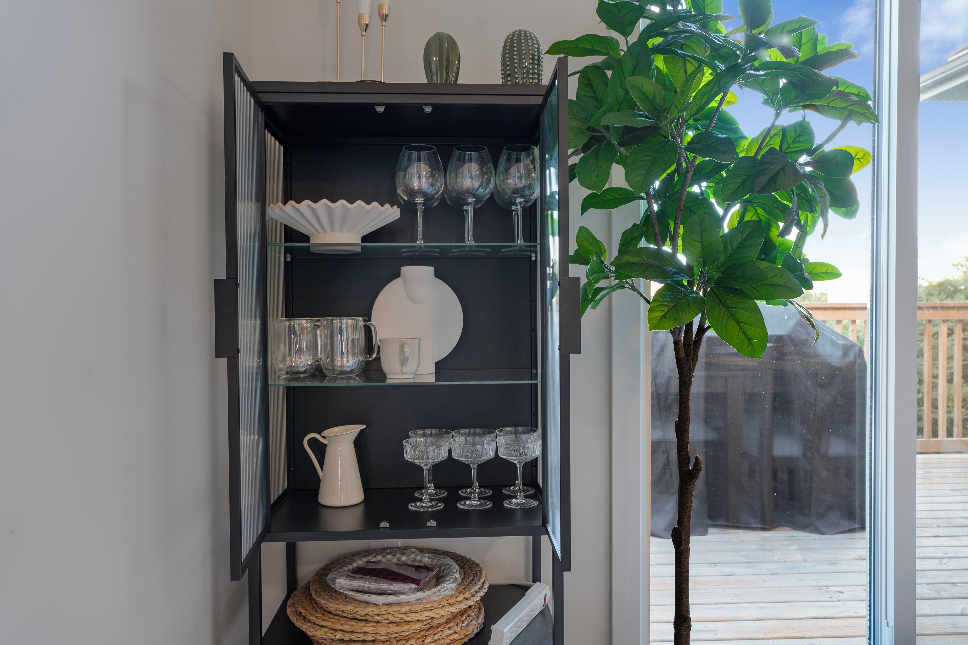 Une étagère noire où sont exposés des objets en verre et des éléments décoratifs, une plante en pot et, en arrière-plan, une vue sur une terrasse en bois.