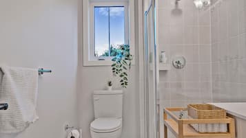 A bathroom with a toilet, a window, a shower, and a wooden shelf with baskets.