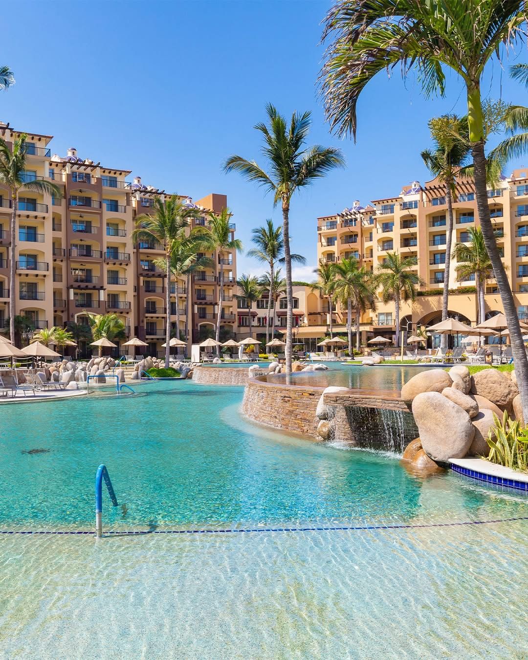 A resort pool area with clear water, palm trees, and a waterfall feature.