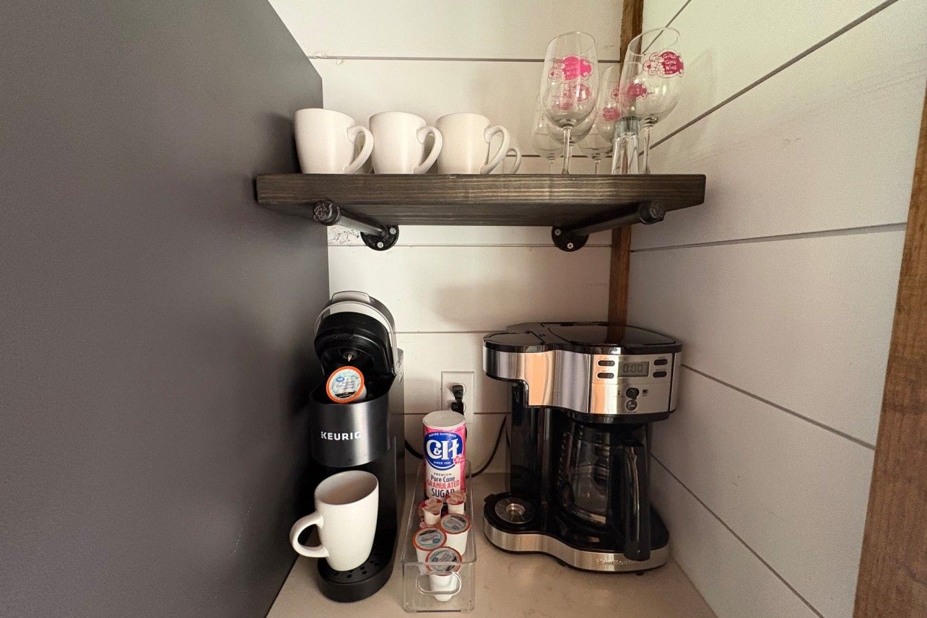 Una zona de cocina con cafetera, taza, vaso de leche y repisa con vasos y copas de vino.