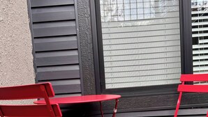 Two red metal folding chairs with a table, positioned outside a building with dark siding and a window with blinds. - Brighton Escape, Spacious 2BR Walk-out Suite (Saskatoon)