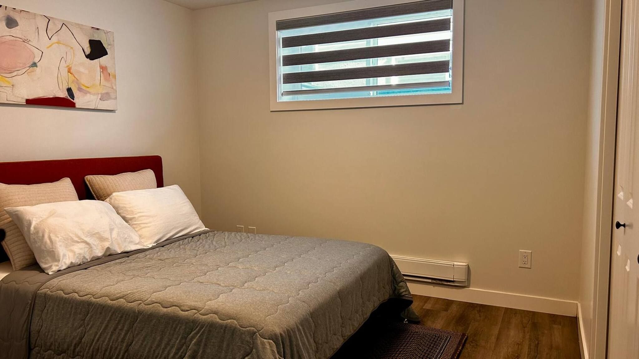 A bedroom with a bed, a window with blinds, a wall-mounted air conditioner, and a partial view of a door.