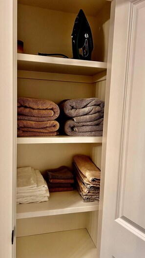 A storage cabinet with shelves holding folded towels and a black iron. - Brighton Escape, Spacious 2BR Walk-out Suite (Saskatoon)