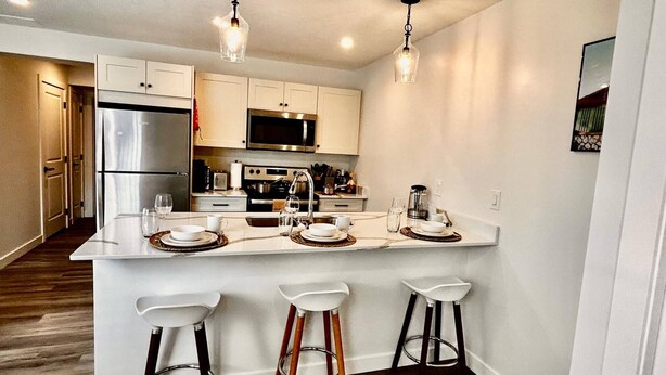 A modern kitchen with white cabinetry, stainless steel appliances, and a bar area with stools. - Brighton Escape, Spacious 2BR Walk-out Suite (Saskatoon)