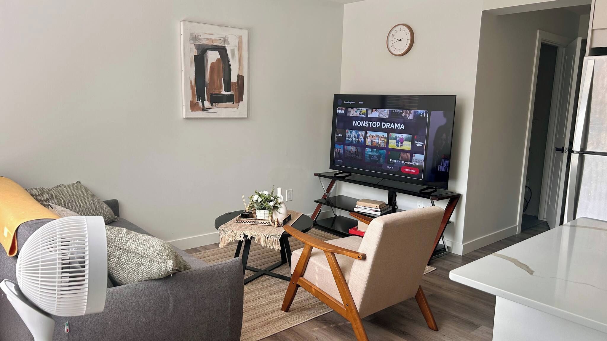 A living room with a flat-screen TV, a sofa, a chair, a small table with a vase, and a clock on the wall.