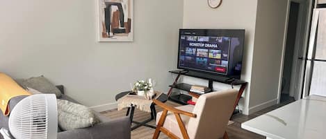 Un salon comprenant un téléviseur à écran plat, un canapé, un fauteuil, une petite table avec un vase et une horloge murale.