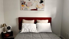 Een slaapkamer met een rood hoofdeinde, twee witte kussens, een grijs gewatteerd sprei, een nachtkastje met een lamp, en een abstract schilderij aan de muur.