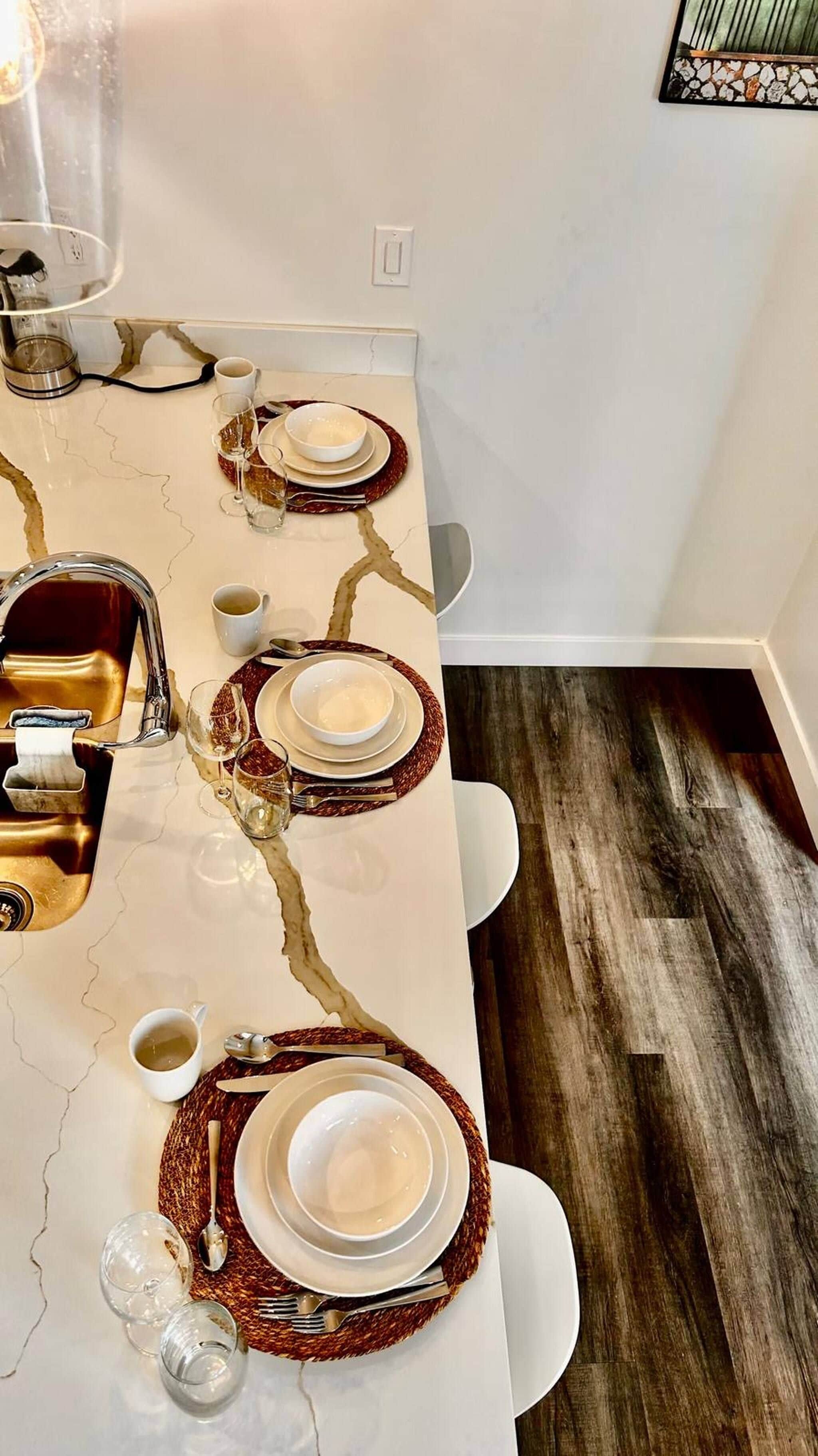 A dining table set with plates, bowls, glasses, and cutlery, with a modern kitchen sink and a light fixture above.