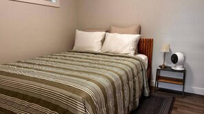 A neatly made bed with a striped bedspread, a bedside table with a lamp and a fan, and a plain wall. - Brighton Escape, Spacious 2BR Walk-out Suite (Saskatoon)