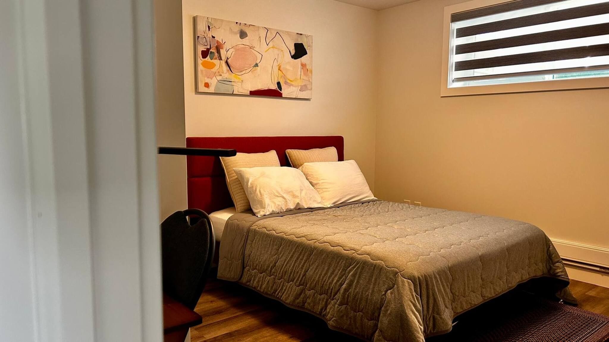 A bedroom with a bed, a chair, a window with blinds, and a wall-mounted abstract painting.
