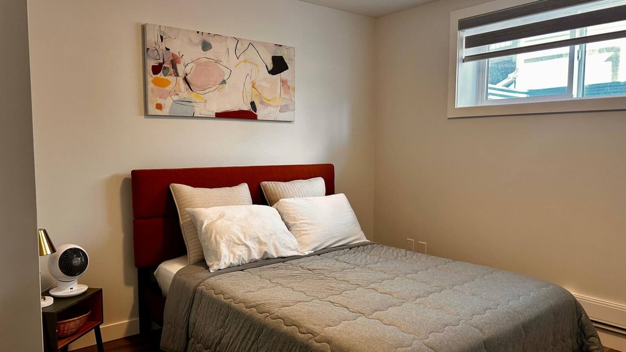 A bedroom with a bed, pillows, a bedside table with a fan, and a window.