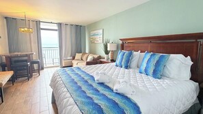 A hotel room with a large bed, a desk, a chair, a lamp, and a view of the sea. - Sunny Oceanfront Studio + pool (Myrtle Beach)