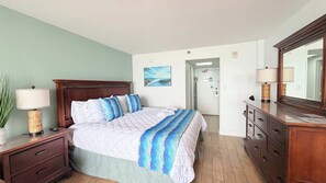 A bedroom with a bed, bedside tables, a dresser, a mirror, a painting, and a door leading to another room. - Sunny Oceanfront Studio + pool (Myrtle Beach)
