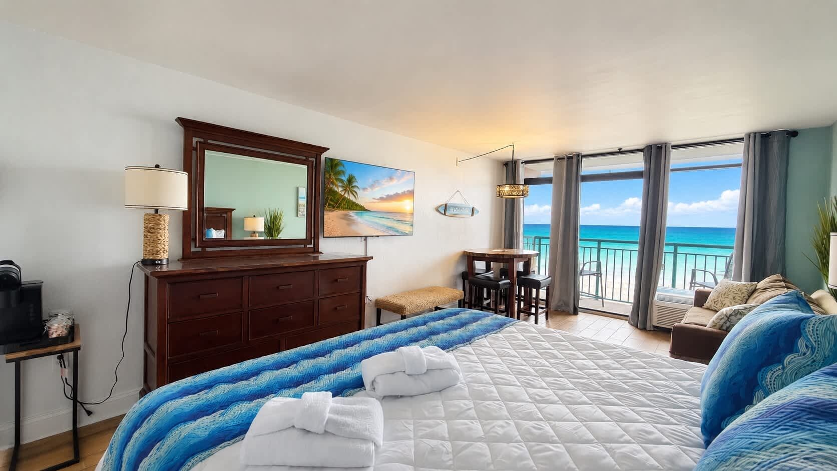 A hotel room with a bed, a desk, a chair, a small table, a sofa, a balcony with a view of the ocean, and a large window.