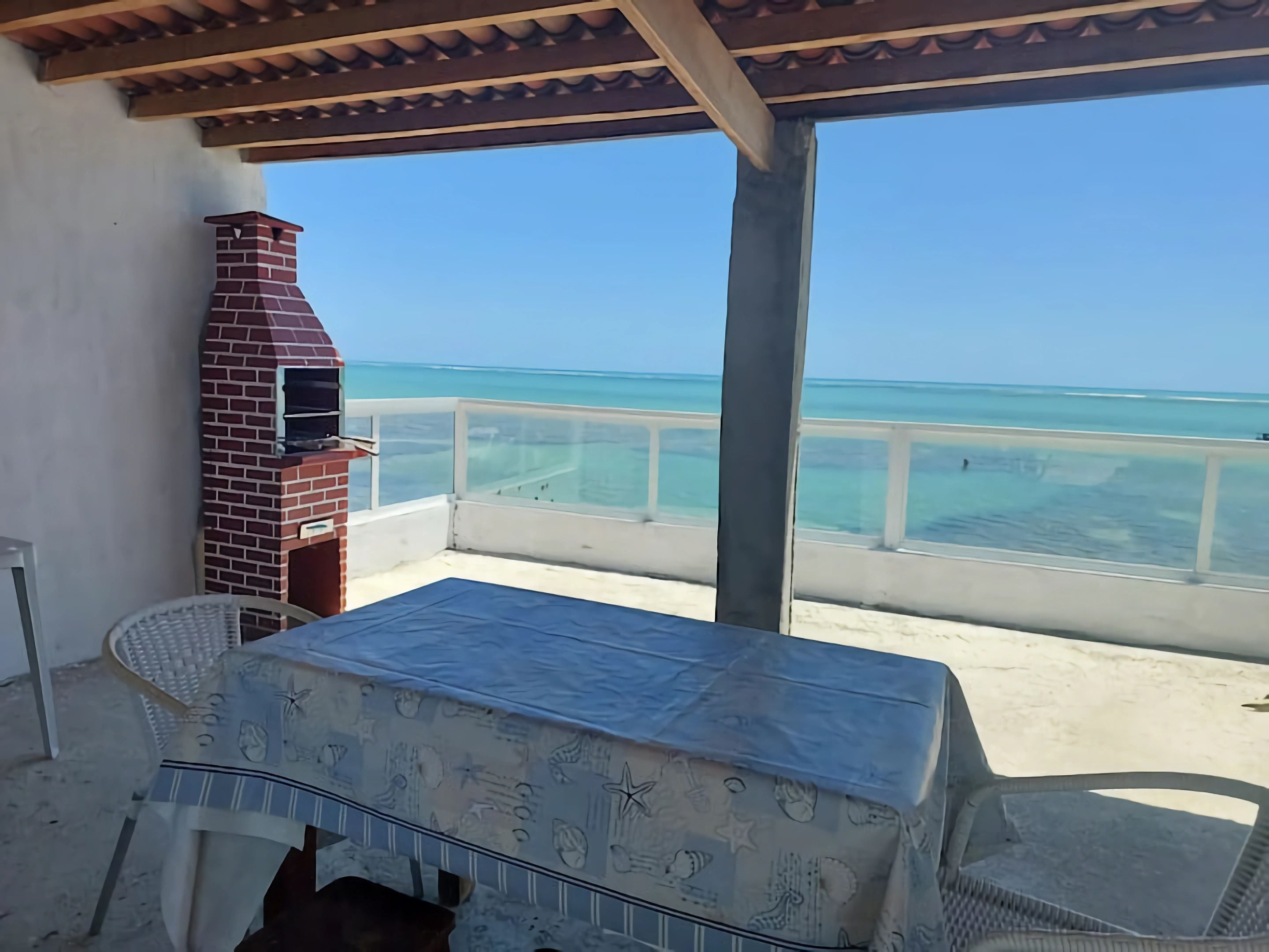 A covered outdoor area with a brick barbecue, a table with a tablecloth, and a view of the ocean.