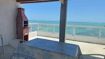 A covered outdoor area with a brick barbecue, a table with a tablecloth, and a view of the ocean.