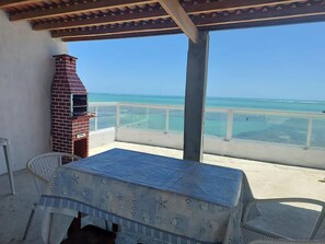 A covered outdoor area with a brick barbecue, a table with a tablecloth, and a view of the ocean.