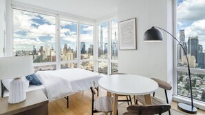 A room with a bed, a round table, chairs, a lamp, and large windows offering a city view.