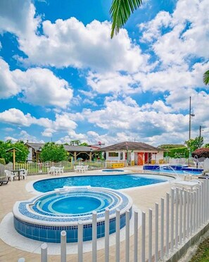 Una piscina con valla perimetral, un área de estar techada y un edificio al fondo.