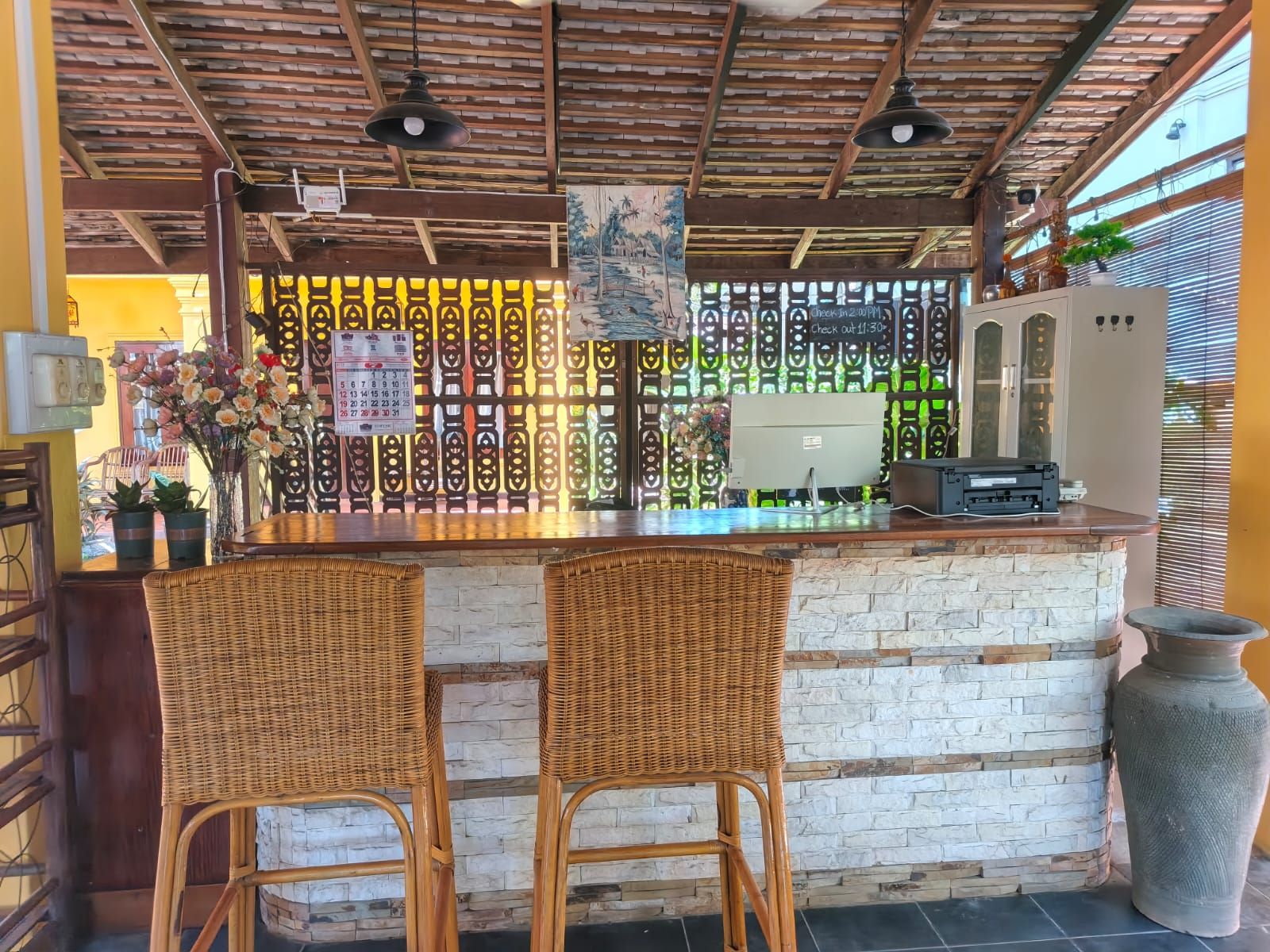Zona de bar con dos sillones de mimbre, una barra con un ordenador y una pared decorativa con un calendario.