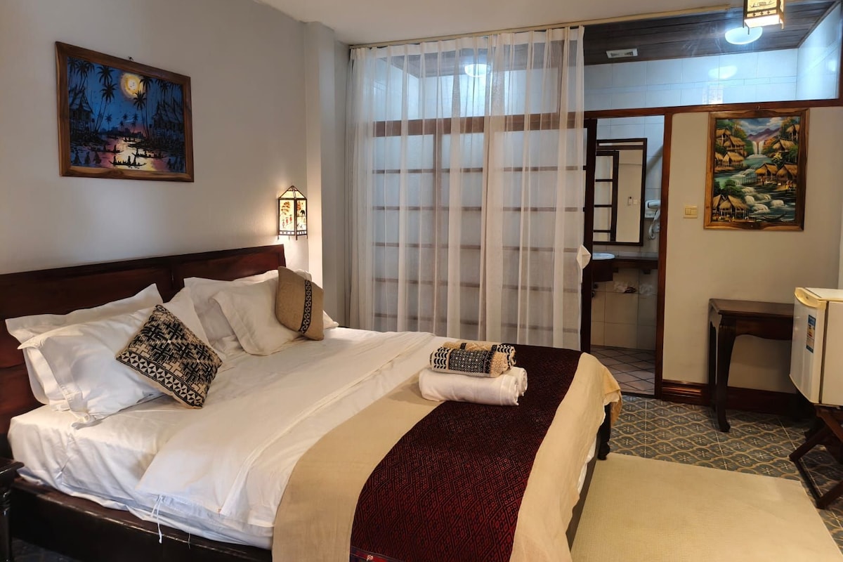Une chambre d’hôtel avec un lit, une table de chevet, une lampe, une fenêtre avec des rideaux légers et un tableau au mur.