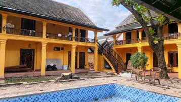 A two-story yellow building with a blue-tiled swimming pool, outdoor seating, and a staircase leading to the upper floor.